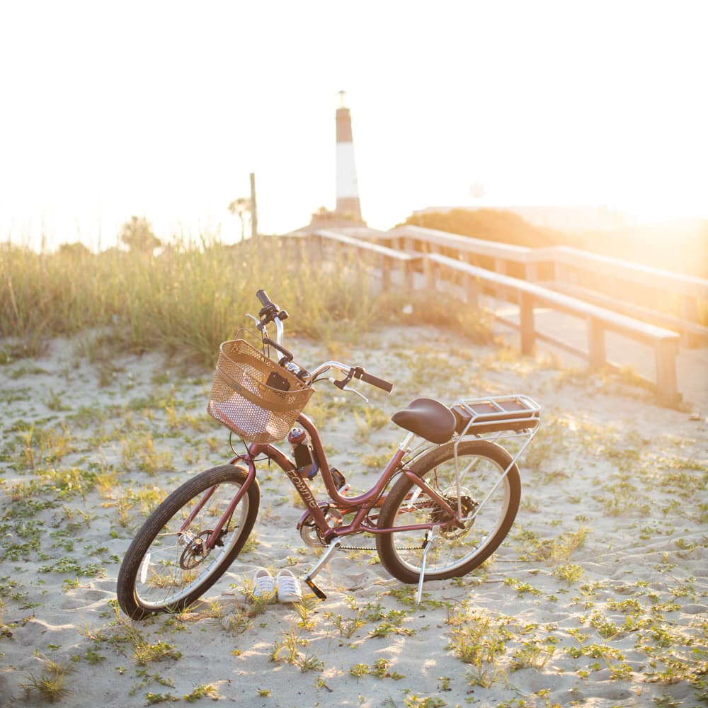 A bicycle with a basket sitting on a grassy beach near a lighthouse.